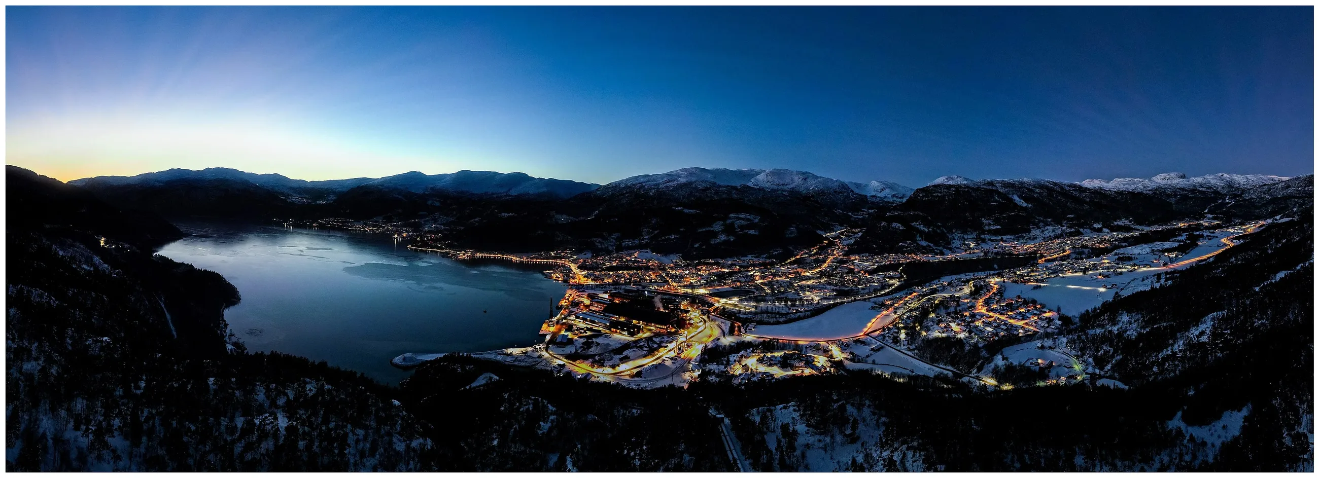 Luftfoto av Sauda sentrum ved fjorden i vinterlys, med snødekte fjell rundt